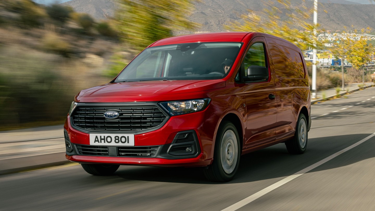 Ford Transit Connect in Maple Red mit Kühlergrill und Scheinwerfern fährt eine Straße entlang. 